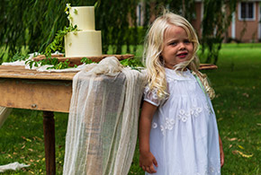 An adorable flower girl poses next to the wedding cake at an outdoor reception on the Frank Phillips Mansion Lawn
